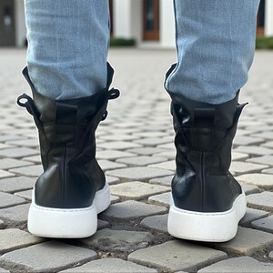 May include: Black leather high-top sneakers with white soles. The boots have a rounded toe and a mid-calf height. The laces are hidden, and the top of the boot is folded over. The sneakers are worn with light blue jeans.
