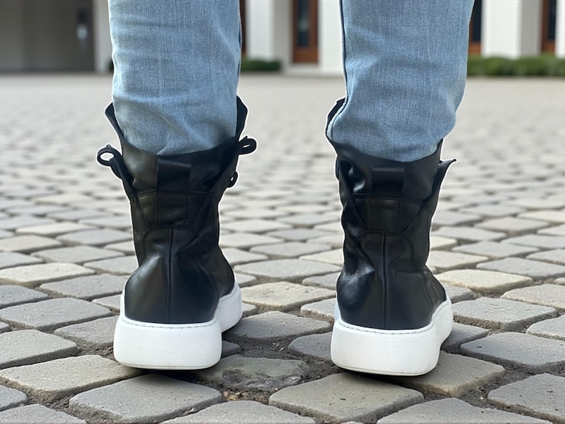 May include: Black leather high-top sneakers with white soles. The boots have a rounded toe and a mid-calf height. The laces are hidden, and the top of the boot is folded over. The sneakers are worn with light blue jeans.