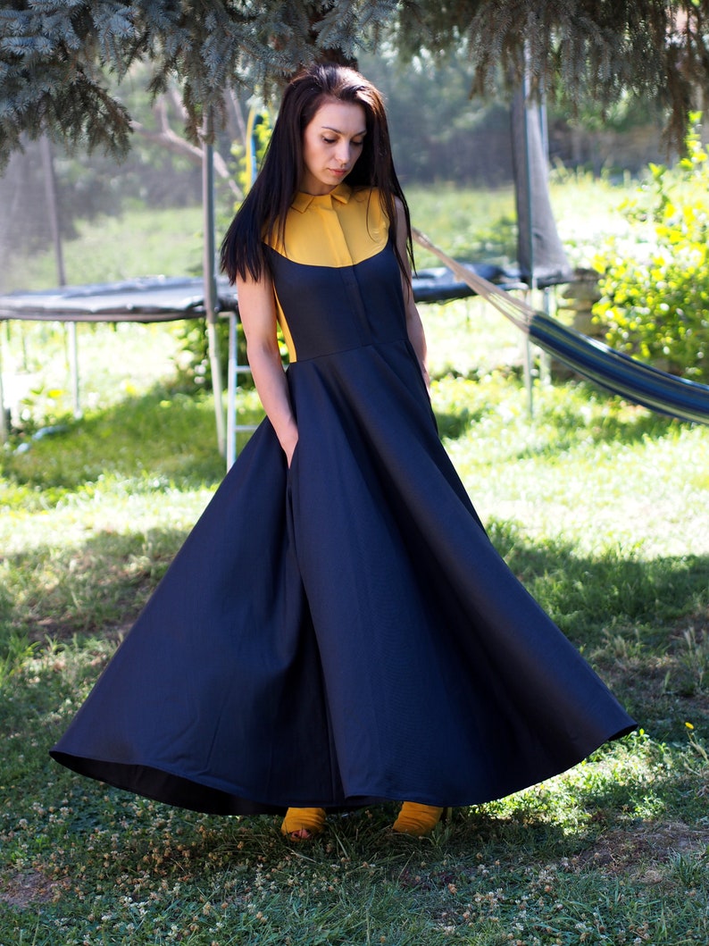 May include: A woman wearing a navy blue dress with a yellow collar and a full skirt. She is standing in a grassy area.