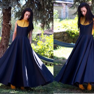 May include: A woman wearing a navy blue maxi dress with a yellow collared shirt underneath. The dress has a full skirt and is sleeveless. The woman is standing in a garden setting.