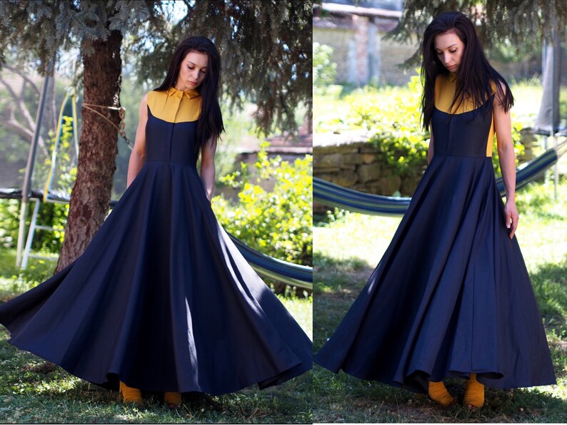 May include: A woman wearing a navy blue maxi dress with a yellow collared shirt underneath. The dress has a full skirt and is sleeveless. The woman is standing in a garden setting.