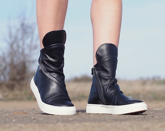 Peut inclure: Bottines en cuir noir avec une semelle en caoutchouc blanche. Les bottes ont un design unique avec un rabat replié à la cheville.