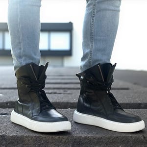 May include: Black high-top sneakers with white soles. The sneakers have a zipper and laces, and are worn with light blue jeans. The shoes have a modern, urban style.