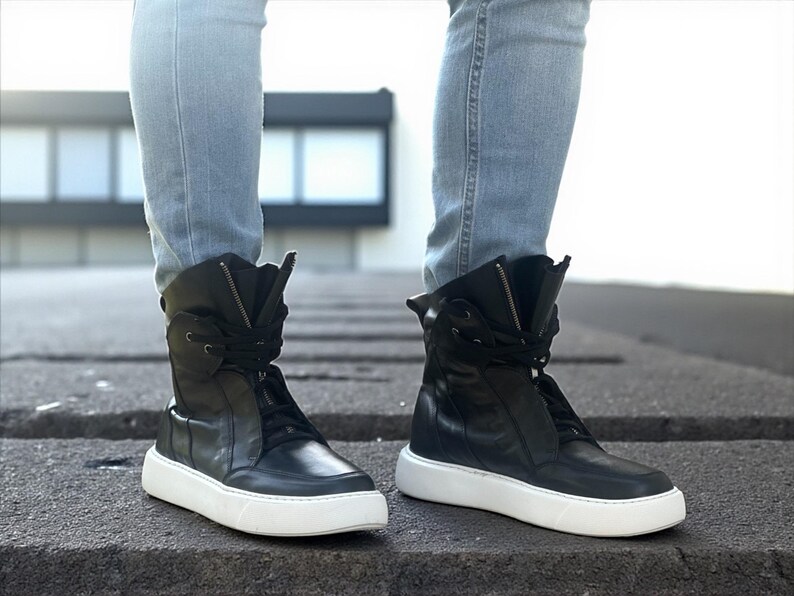 May include: Black high-top sneakers with white soles. The sneakers have a zipper and laces, and are worn with light blue jeans. The shoes have a modern, urban style.