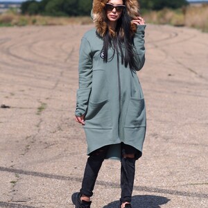 May include: A long, sage green hooded sweatshirt with a faux fur hood and a zipper closure. The sweatshirt has a high-low hemline and large pockets. The model is wearing black jeans and black sneakers.