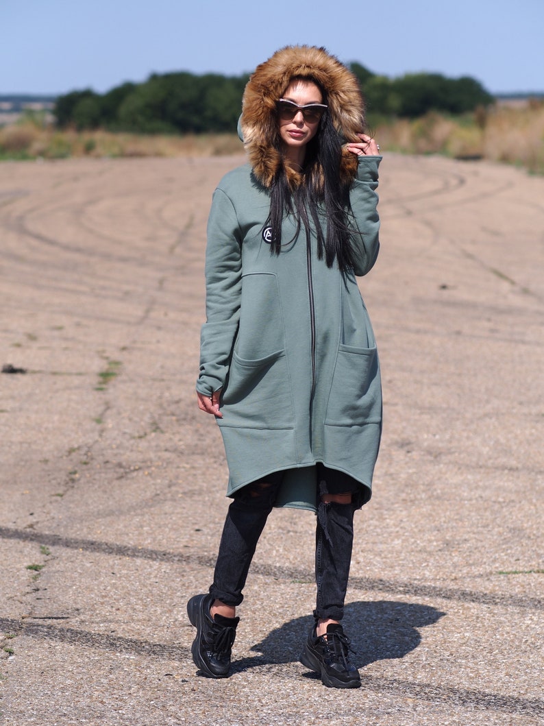 May include: A long, sage green hooded sweatshirt with a faux fur hood and a zipper closure. The sweatshirt has a high-low hemline and large pockets. The model is wearing black jeans and black sneakers.