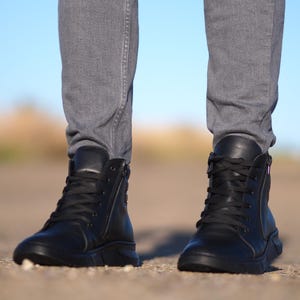 May include: Black leather high-top boots with laces and side zippers. The boots have a thick sole and are paired with gray denim pants. The image is taken from a low angle, showing the boots in detail.
