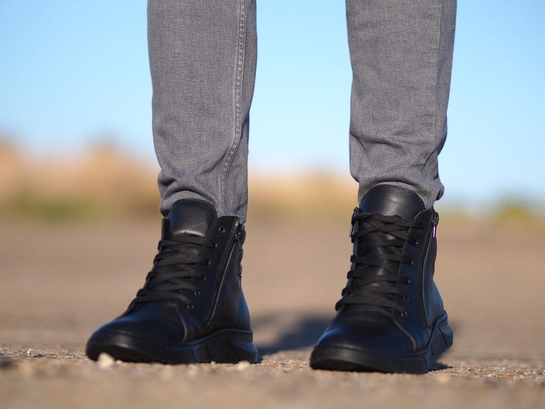 May include: Black leather high-top boots with laces and side zippers. The boots have a thick sole and are paired with gray denim pants. The image is taken from a low angle, showing the boots in detail.