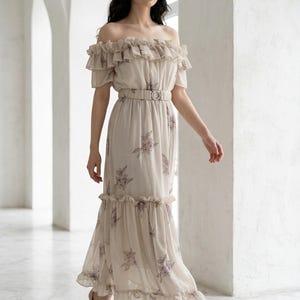 May include: A beige off-the-shoulder maxi dress with a floral pattern and a belted waist. The dress features ruffled detailing at the neckline and hem. The model is wearing nude high heels.