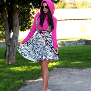 May include: A woman wearing a pink hooded sweatshirt and a black and white patterned skirt with a black belt that says "ICON GIRL" repeated. She is wearing sunglasses and white sneakers.
