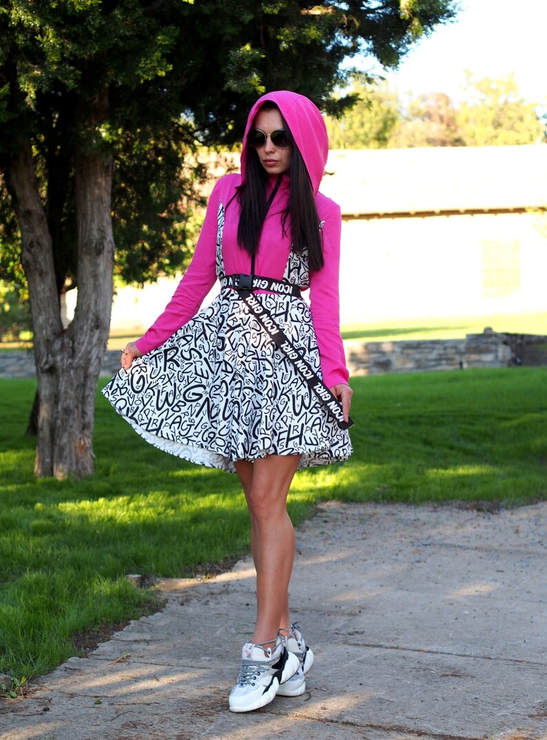 May include: A woman wearing a pink hooded sweatshirt and a black and white patterned skirt with a black belt that says "ICON GIRL" repeated. She is wearing sunglasses and white sneakers.