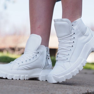 Puede incluir: Un par de zapatillas blancas de caña alta con suela gruesa y cordones. Las zapatillas tienen una cremallera en el lateral.