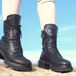 K&ouml;nnte beinhalten: Ein Paar schwarze Lederstiefel mit Schn&uuml;rung und einem dicken Absatz. Die Stiefel haben ein Schlangenhautmuster an Zehen und Ferse.