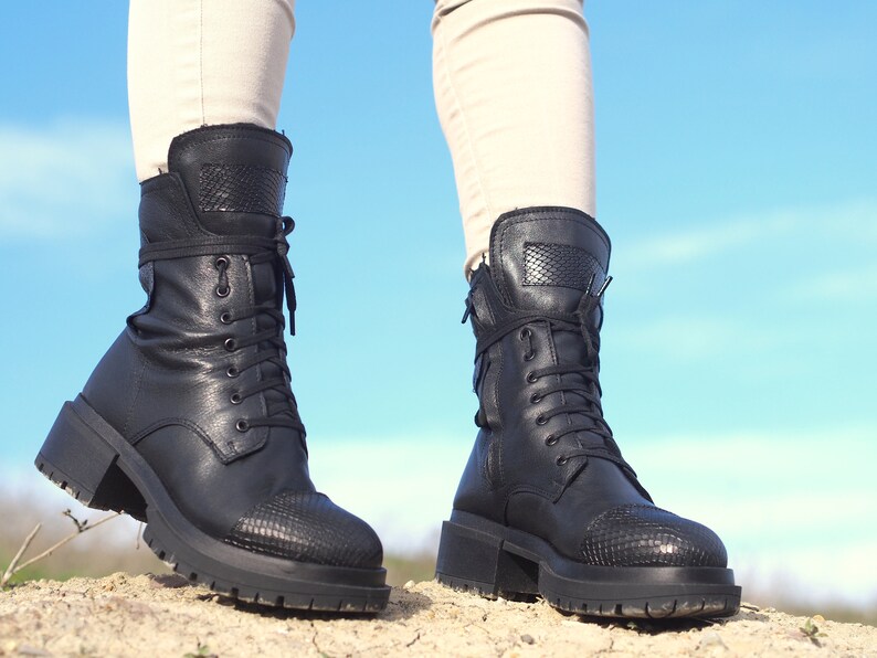 K&ouml;nnte beinhalten: Ein Paar schwarze Lederstiefel mit Schn&uuml;rung und einem dicken Absatz. Die Stiefel haben ein Schlangenhautmuster an Zehen und Ferse.