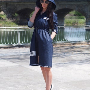 May include: A dark blue denim dress with a hood and long sleeves. The dress has a drawstring waist and white text on the bottom hem. The model is wearing sunglasses and white sneakers.