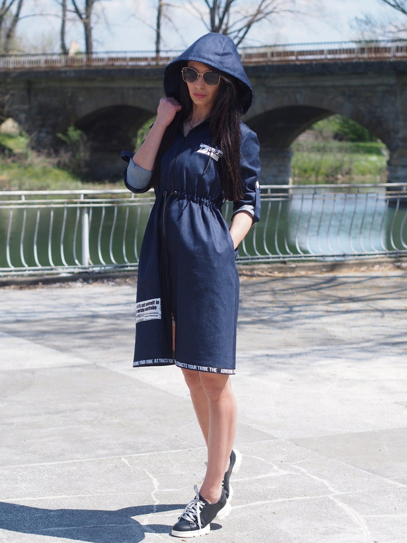 May include: A dark blue denim dress with a hood and long sleeves. The dress has a drawstring waist and white text on the bottom hem. The model is wearing sunglasses and white sneakers.
