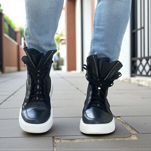 May include: Black high-top sneakers with white soles and a zipper detail. The sneakers have laces and a folded-over design at the top. The shoes are worn with light blue jeans.