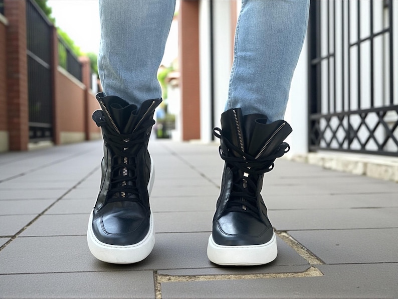 May include: Black high-top sneakers with white soles and a zipper detail. The sneakers have laces and a folded-over design at the top. The shoes are worn with light blue jeans.