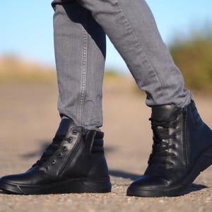 May include: Black leather high-top sneakers with side zippers and lace-up closures. The shoes have a thick, textured sole and are worn with gray jeans. The image is taken outdoors on a sunny day.