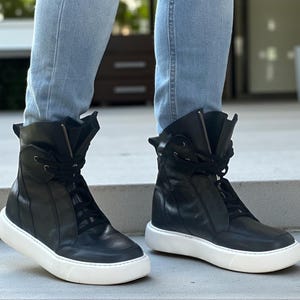 May include: A pair of black high-top sneakers with white soles. The sneakers have laces and zippers, and are made of leather. The shoes are worn with light blue jeans.