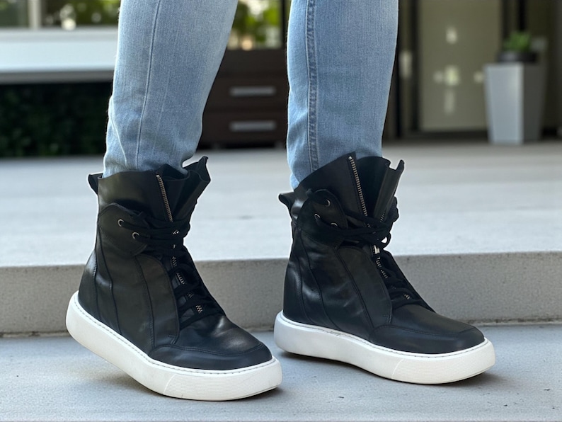 May include: A pair of black high-top sneakers with white soles. The sneakers have laces and zippers, and are made of leather. The shoes are worn with light blue jeans.