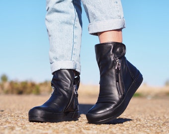 Peut inclure: Une paire de bottines en cuir noir à fermeture éclair latérale. Les bottes ont une semelle épaisse et sont portées avec un jean bleu retroussé.