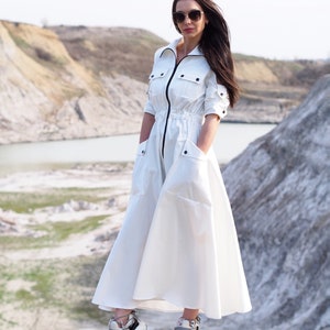 May include: A white long-sleeved dress with a zipper down the front and pockets on the sides. The dress has a cinched waist and a flowing skirt. The model is wearing the dress with white sneakers and sunglasses.