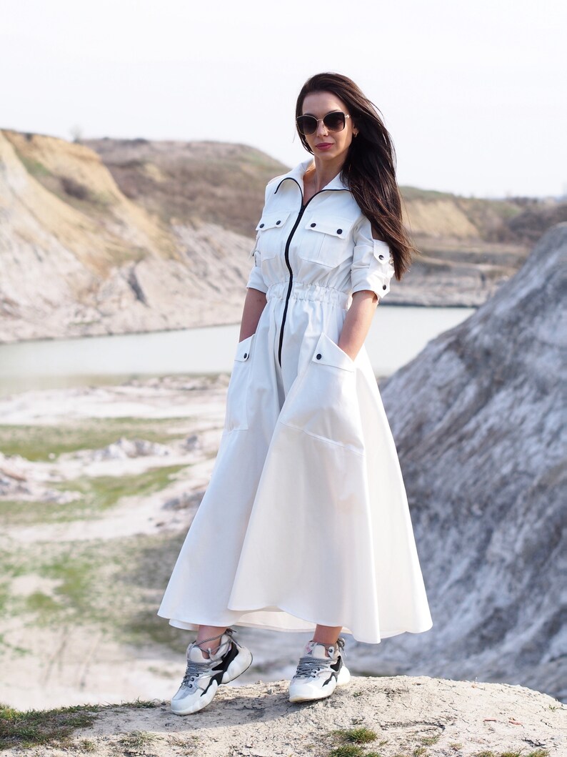 May include: A white long-sleeved dress with a zipper down the front and pockets on the sides. The dress has a cinched waist and a flowing skirt. The model is wearing the dress with white sneakers and sunglasses.