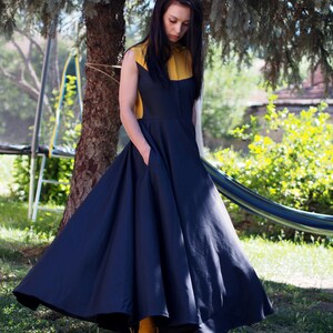 May include: A woman wearing a navy blue and yellow dress with a full skirt. She is standing in a grassy area with a tree in the background.