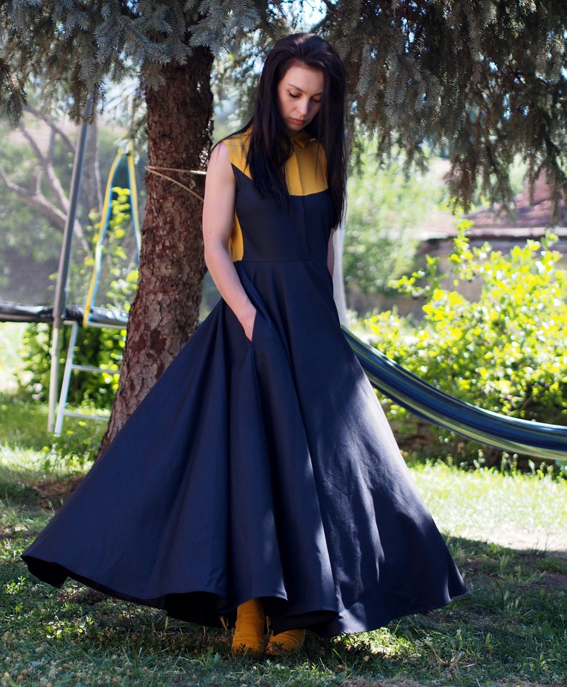 May include: A woman wearing a navy blue and yellow dress with a full skirt. She is standing in a grassy area with a tree in the background.