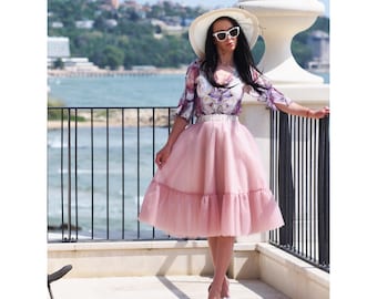 May include: A woman wearing a pink tulle skirt with a floral print blouse and a white hat. She is standing on a balcony overlooking the ocean.