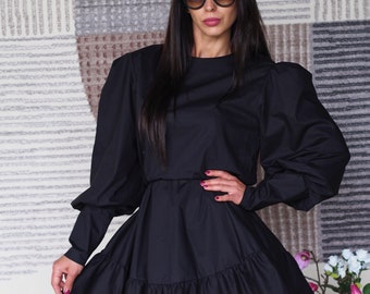 May include: A black dress with a ruffled skirt and long sleeves. The dress has a round neckline and is worn with a black cap and sunglasses.