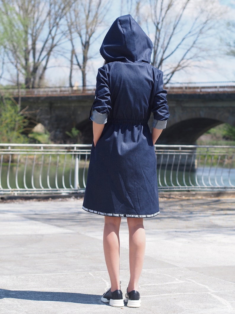 May include: A dark blue denim hooded jacket with a drawstring waist. The jacket has white text on the hem that reads "Your vibe attracts your tribe. The adventure begins your vibe."