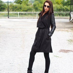 May include: A black trench coat with leather sleeves. The coat has a belt and a collar. The model is wearing black tights and black ankle boots.
