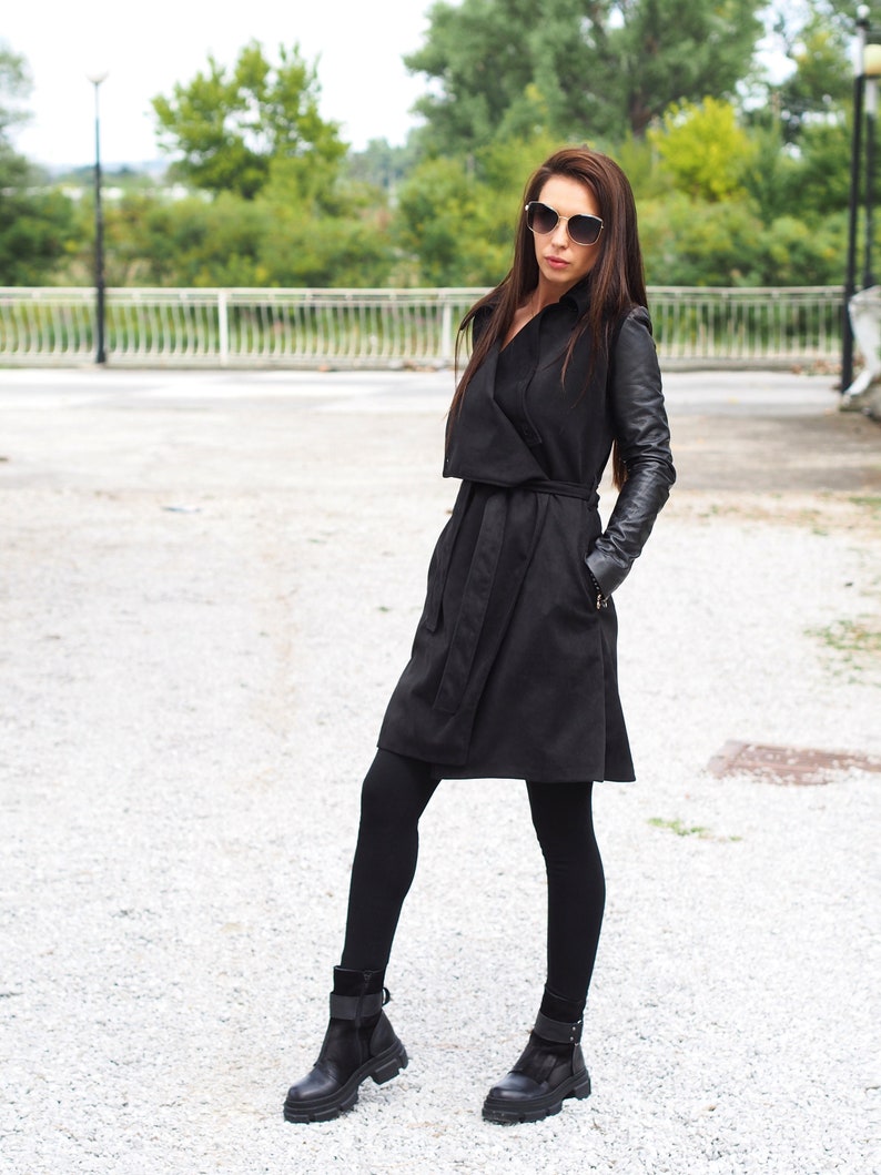 May include: A black trench coat with leather sleeves. The coat has a belt and a collar. The model is wearing black tights and black ankle boots.