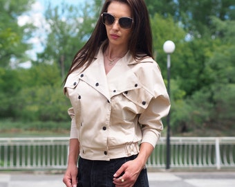Puede incluir: Una mujer con una chaqueta beige de doble botonadura y pantalones cortos de mezclilla negros. Lleva gafas de sol y un collar de oro.