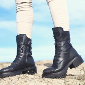 K&ouml;nnte beinhalten: Ein Paar schwarze Lederstiefel mit Schn&uuml;rung und dicken Sohlen. Die Stiefel haben einen seitlichen Rei&szlig;verschluss und werden von einer Person getragen, die auf einem Feldweg steht.