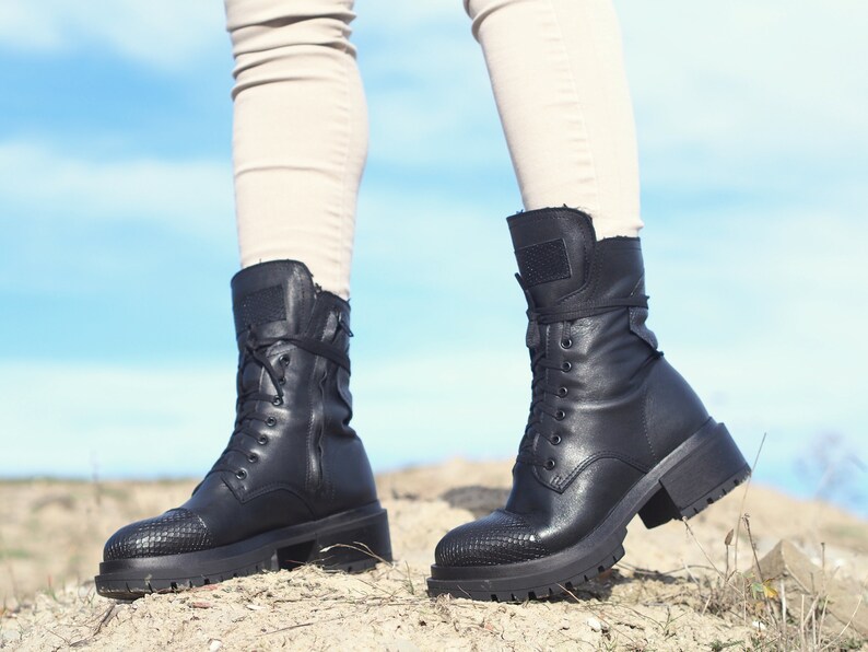 K&ouml;nnte beinhalten: Ein Paar schwarze Lederstiefel mit Schn&uuml;rung und dicken Sohlen. Die Stiefel haben einen seitlichen Rei&szlig;verschluss und werden von einer Person getragen, die auf einem Feldweg steht.