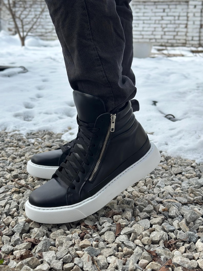 May include: Black high-top trainers with a white platform sole and a zip detail on the side. The trainers are made of leather and have a lace-up closure.