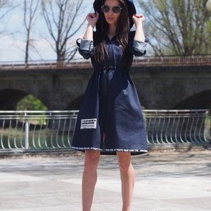 May include: A dark blue denim dress with a hood and a zipper closure. The dress has a drawstring at the waist and features white text on the bottom hem. The model is wearing black sneakers.
