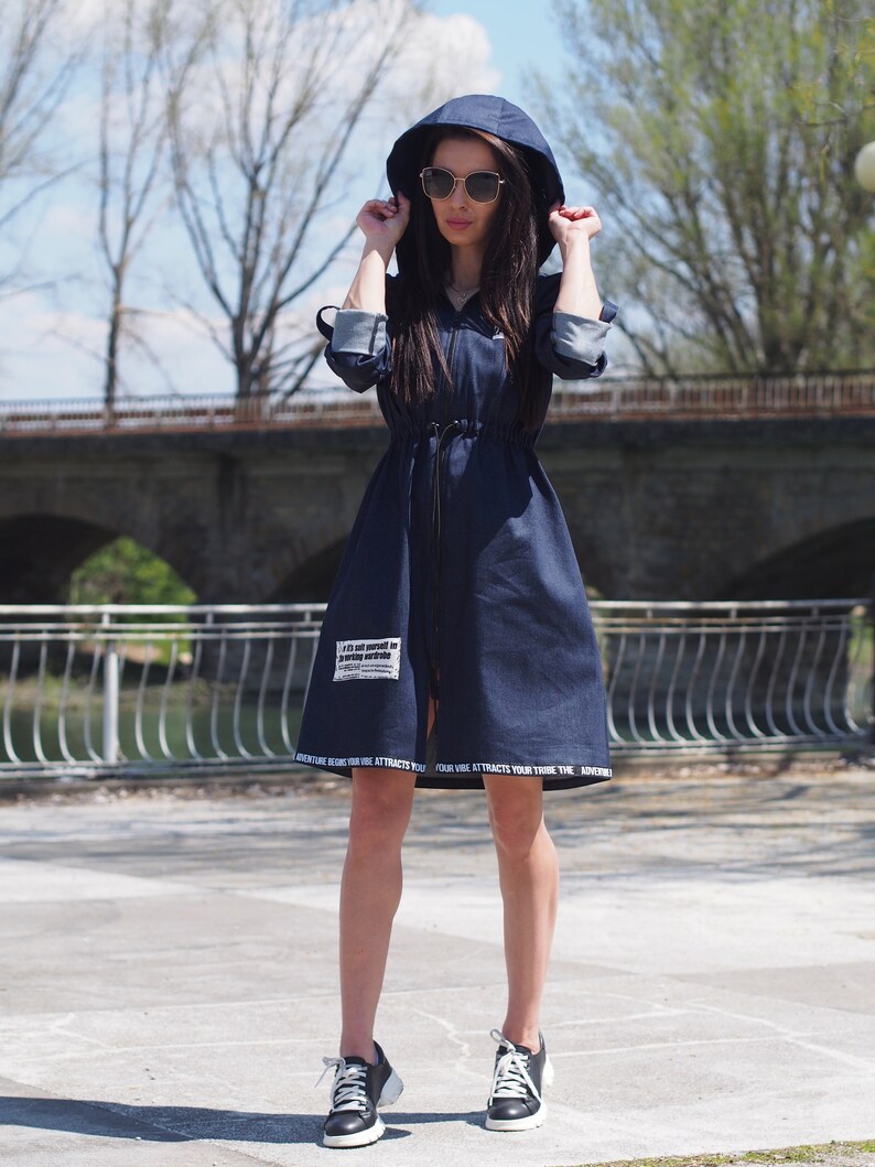 May include: A dark blue denim dress with a hood and a zipper closure. The dress has a drawstring at the waist and features white text on the bottom hem. The model is wearing black sneakers.
