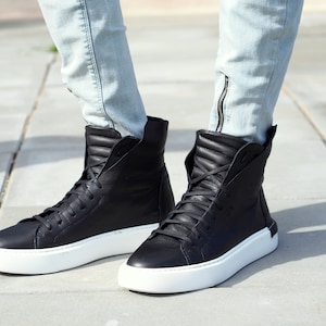 May include: Black leather high-top sneakers with white soles. The sneakers have a zipper detail on the side and are worn with light blue jeans.