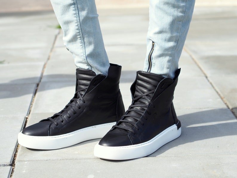 May include: Black leather high-top sneakers with white soles. The sneakers have a zipper detail on the side and are worn with light blue jeans.