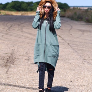 May include: A long, teal green hooded sweatshirt with a brown faux fur hood. The sweatshirt has a zipper closure and a large pocket on the front. The model is wearing black ripped jeans and black sneakers.