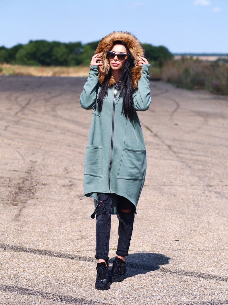 May include: A long, teal green hooded sweatshirt with a brown faux fur hood. The sweatshirt has a zipper closure and a large pocket on the front. The model is wearing black ripped jeans and black sneakers.