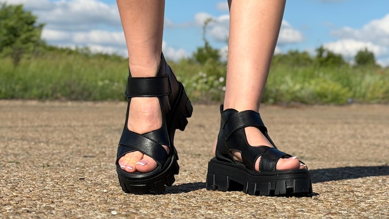 Pu&ograve; includere: Sandali neri con plateau e suola spessa. I sandali hanno un design a cinturini con cinturino alla caviglia regolabile. L'immagine mostra i sandali ai piedi di una persona, con uno sfondo di strada e vegetazione.