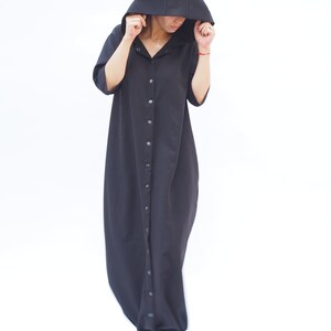 Puede incluir: Un vestido maxi negro con capucha, cierre de botones en la parte delantera y mangas largas. El vestido tiene un corte holgado y una silueta fluida.