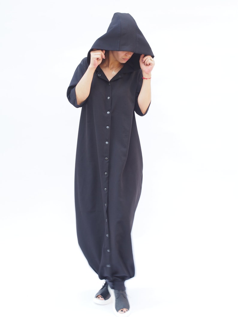 Puede incluir: Un vestido maxi negro con capucha, cierre de botones en la parte delantera y mangas largas. El vestido tiene un corte holgado y una silueta fluida.