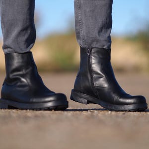 May include: Black leather ankle boots with a side zipper and a rounded toe. The boots have a low, chunky heel and are worn with gray denim pants. The boots are approximately 6 inches in height.