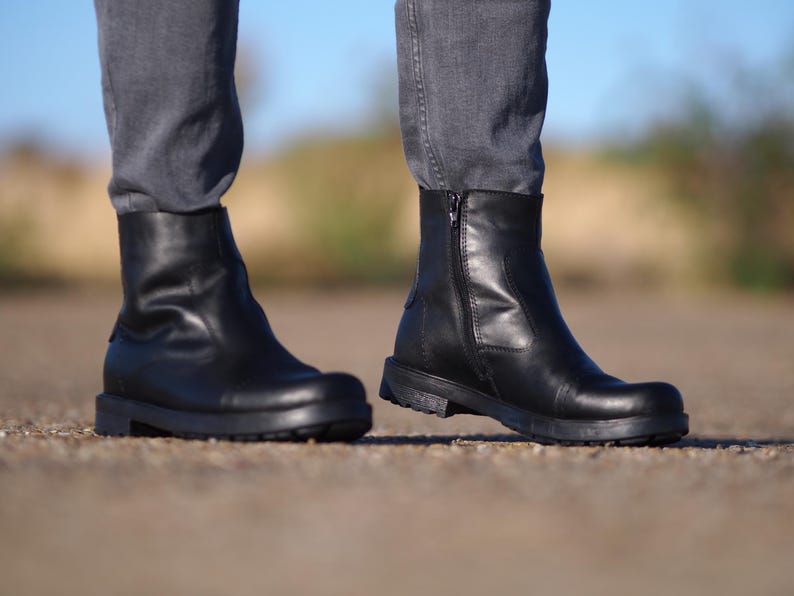 May include: Black leather ankle boots with a side zipper and a rounded toe. The boots have a low, chunky heel and are worn with gray denim pants. The boots are approximately 6 inches in height.